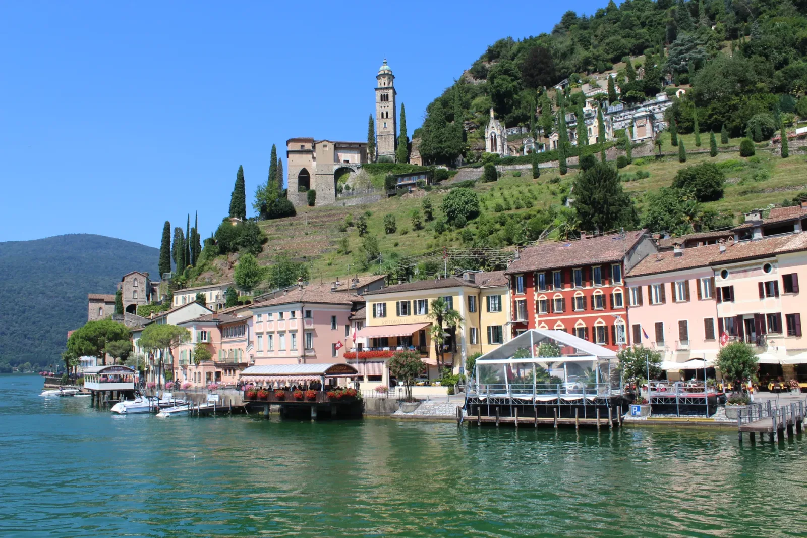 Vista de Morcote às margens do Lago Lugano, com casas históricas e montanhas ao fundo.